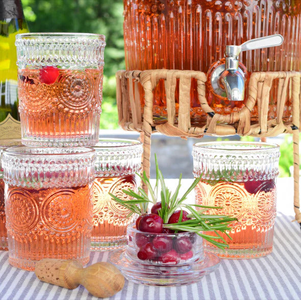 Clear vintage-style embossed glass tumbler filled with pink cocktail next to a matching glass beverage dispenser on a woven rattan stand.