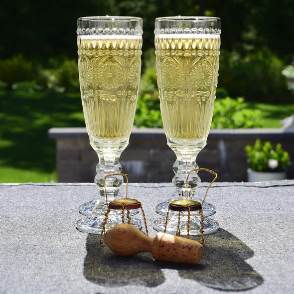 Two vintage-style clear glass champagne flutes filled with bubbly wine, standing behind a wooden wine stopper and metal wire cages on a gray fabric surface.
