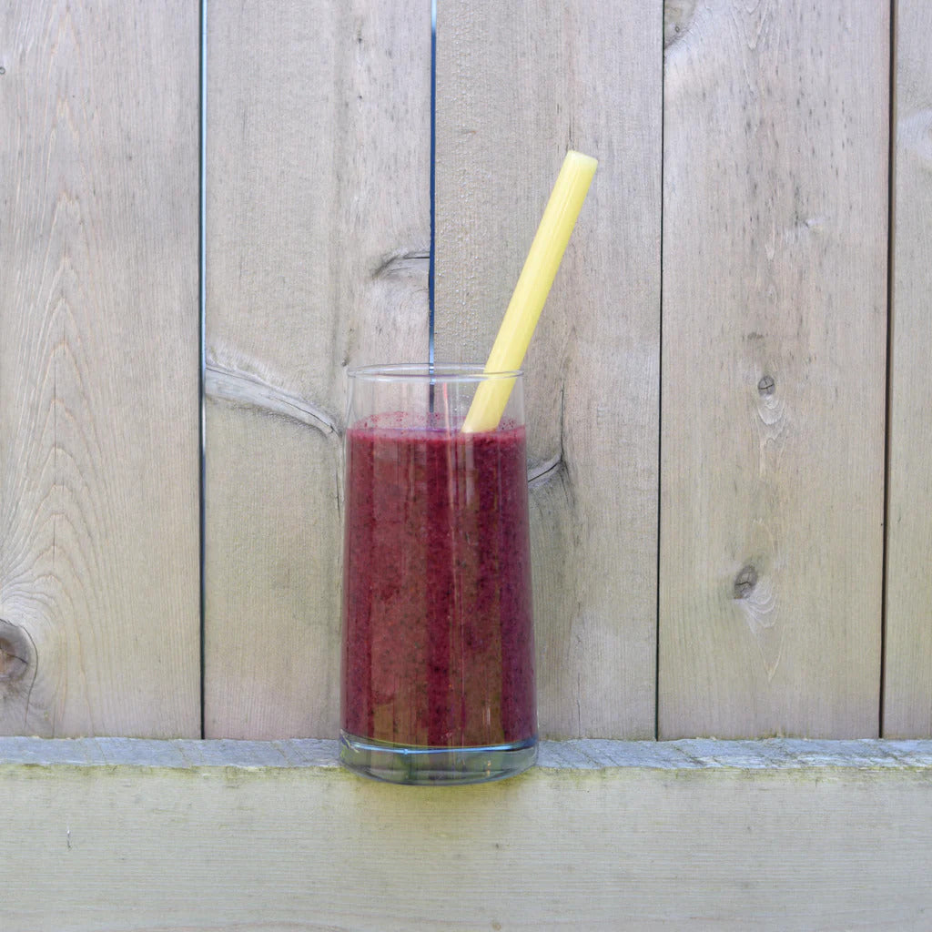 A reusable amber-colored borosilicate glass straw resting in a tall glass of green smoothie against a light rustic wood fence backdrop.