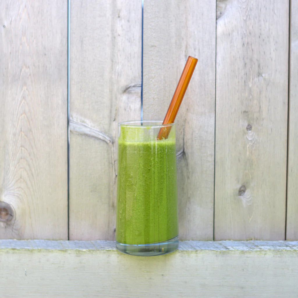 A reusable amber-colored borosilicate glass straw resting in a tall glass of green smoothie against a light rustic wood fence backdrop.