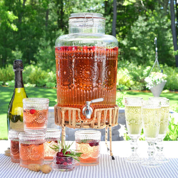 Elegant embossed glass beverage dispenser filled with iced tea and fruit garnish, resting on a natural woven rattan stand on an outdoor dining table.