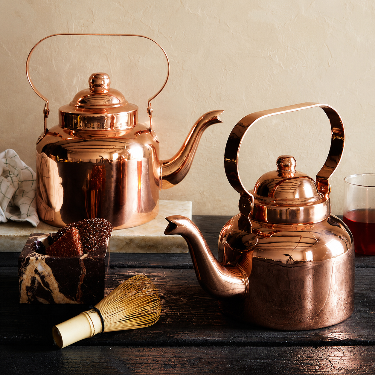 Polished pure copper tea kettle with a curved overhead handle and matching dome lid, resting on a textured cream linen fabric background.